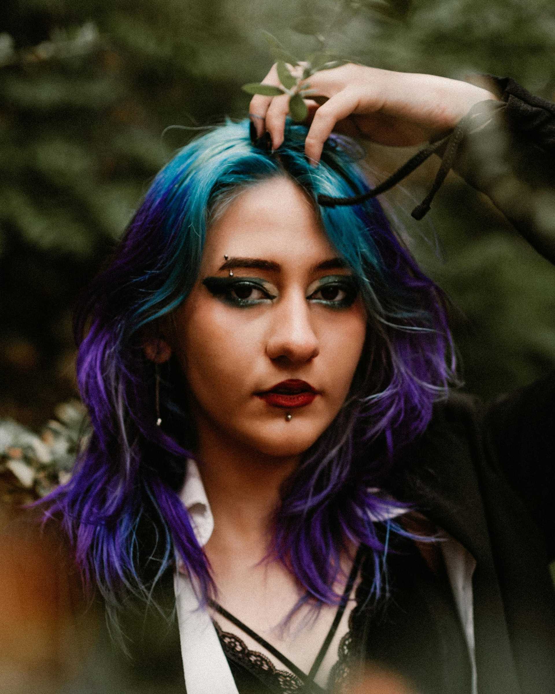 Gothic style woman with vibrant blue and purple hair, intense makeup, and foliage background.